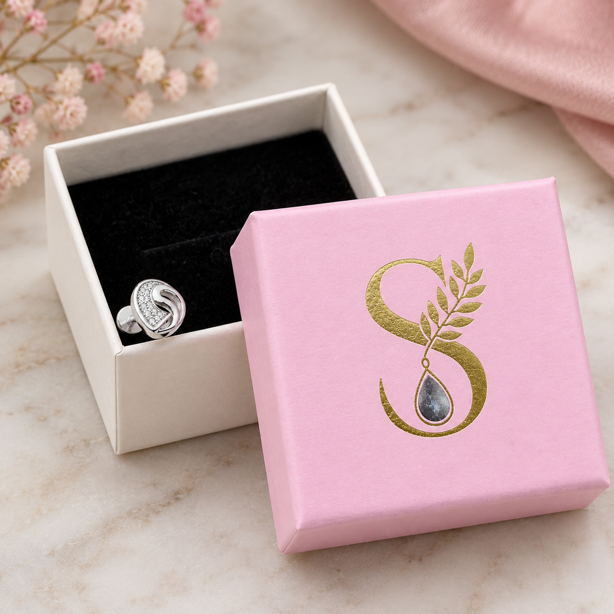 A silver stud earring resting on a pale pink and white jewelry box with a gold monogram.