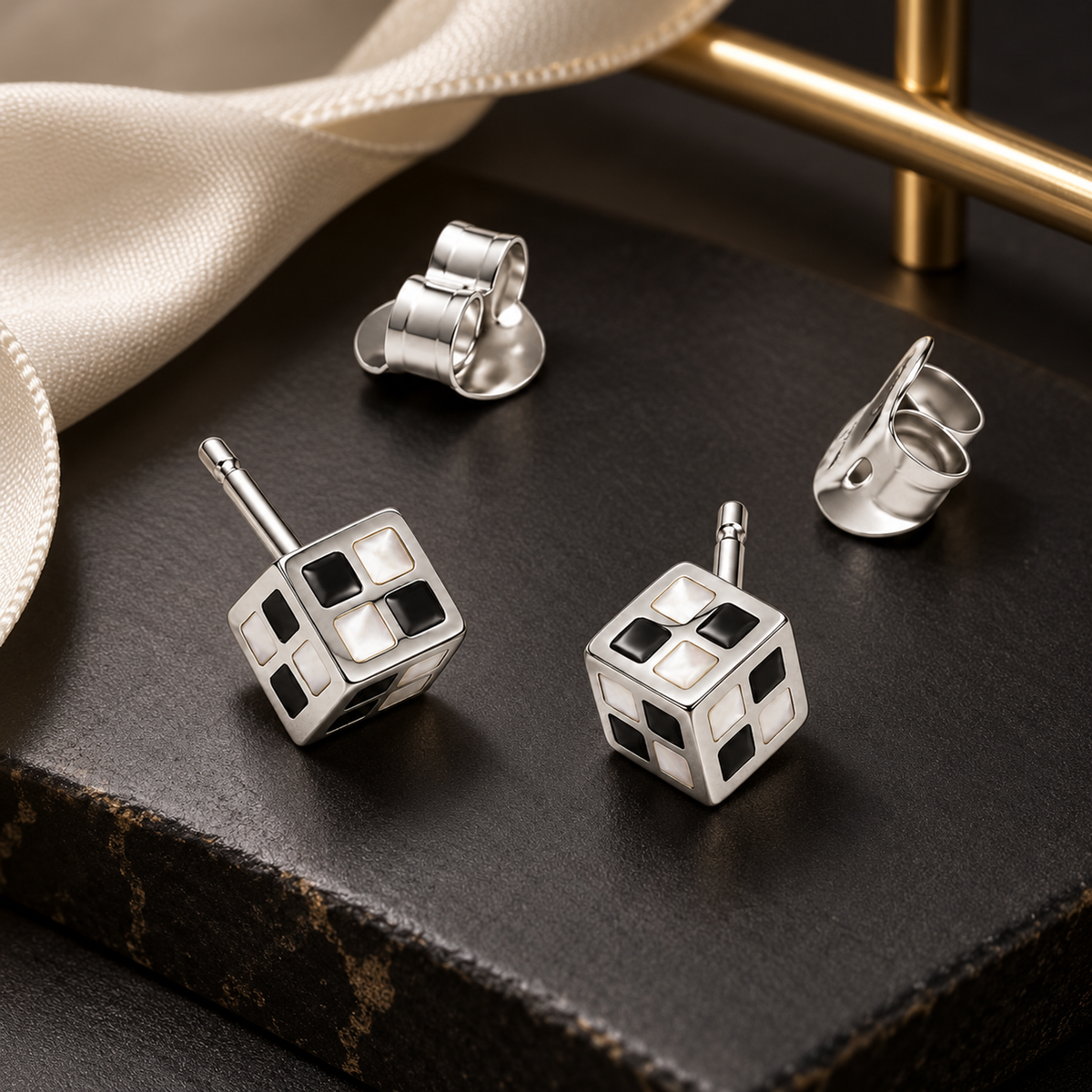 Checkerboard cube stud earrings resting on a matte black ceramic block beside ivory ribbon.
