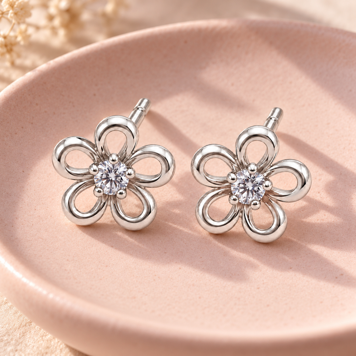 Silver flower stud earrings resting on a pale blush pink ceramic dish.