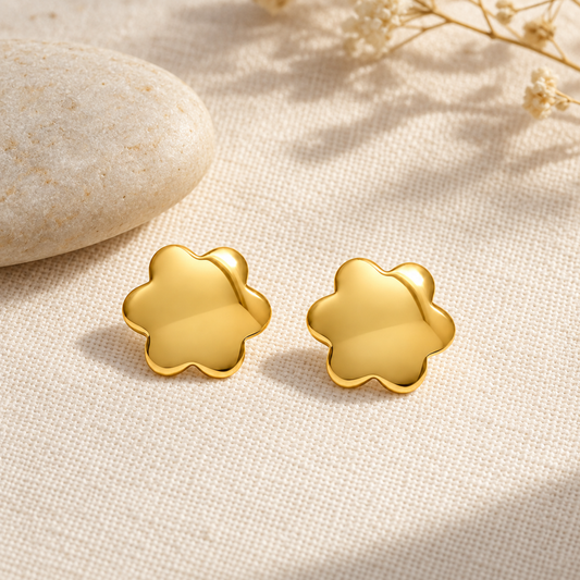 Gold blossom stud earrings on textured ivory linen beside a smooth pebble.
