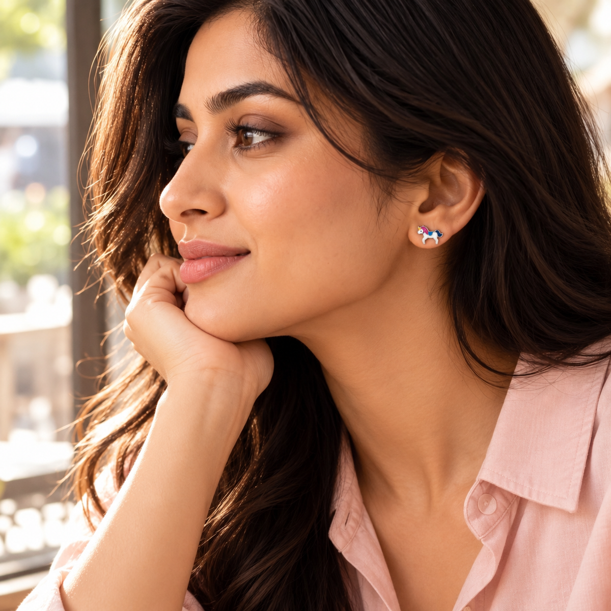 Woman in profile smiling gently by a window, wearing a small silver unicorn stud earring.