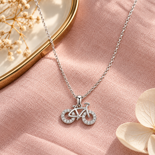Silver bicycle necklace styled on blush pink linen alongside a warm gold tray.