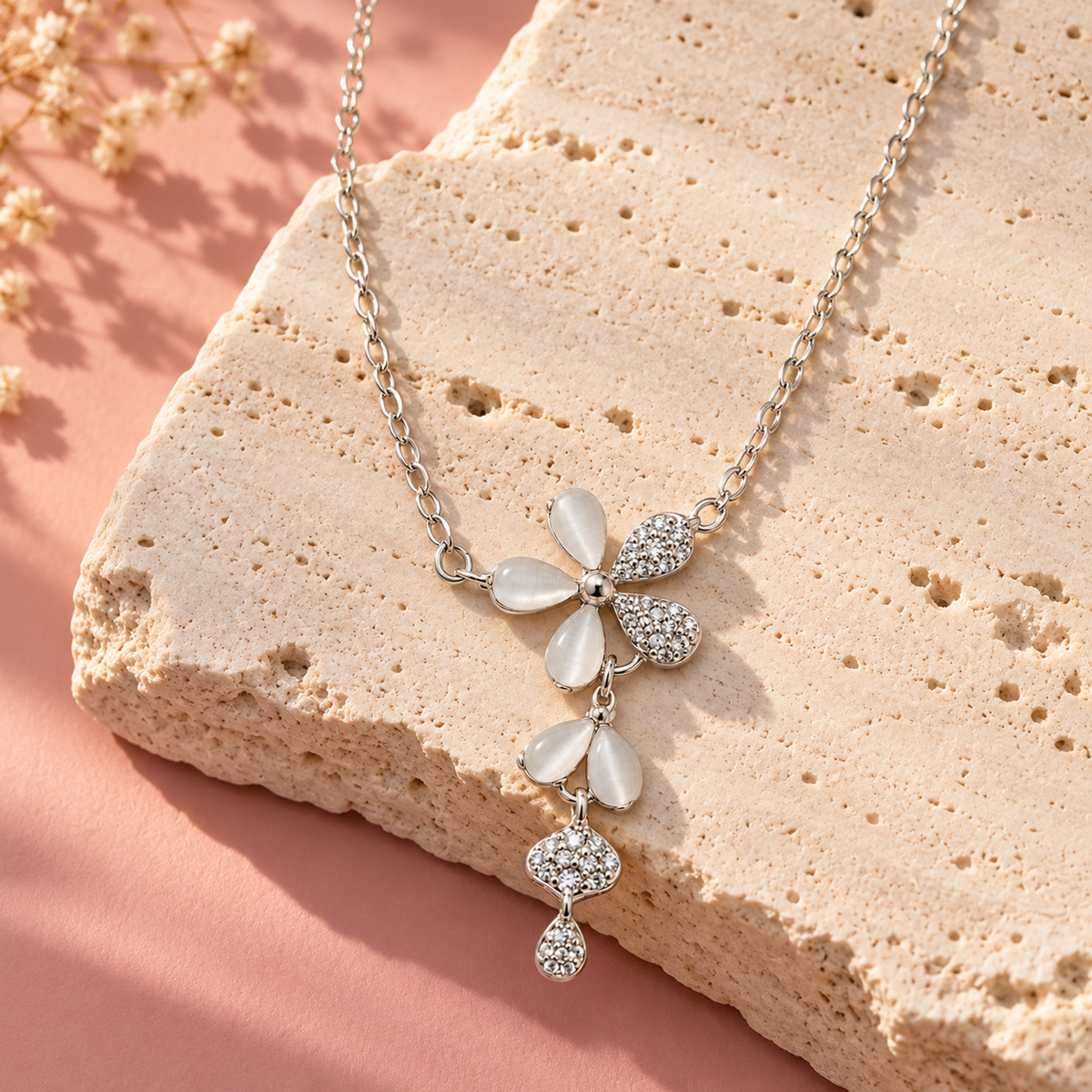 A silver flower necklace on a textured ivory stone against a soft blush pink backdrop in warm morning light.