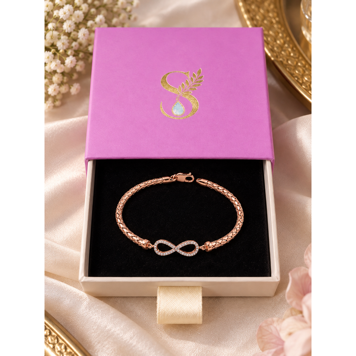 Rose gold infinity bracelet displayed inside open Silvanya magenta drawer gift box on black velvet.