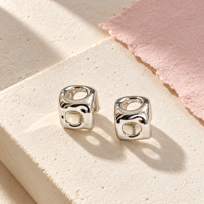 Silver cube stud earrings resting on a pale ivory plaster block with soft geometric shadows.