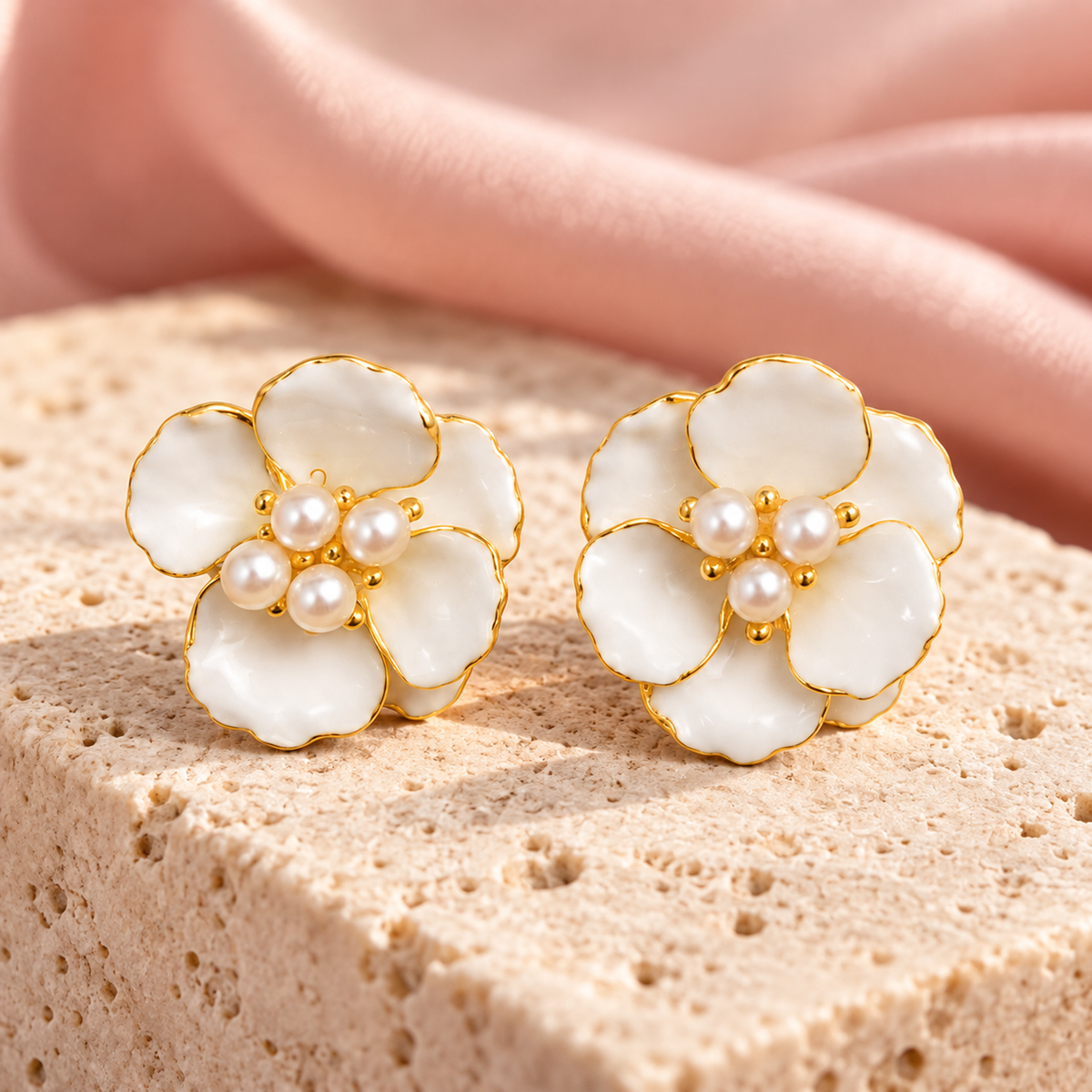 Pair of floral stud earrings resting on a textured ivory travertine stone with soft blush pink silk draped behind.