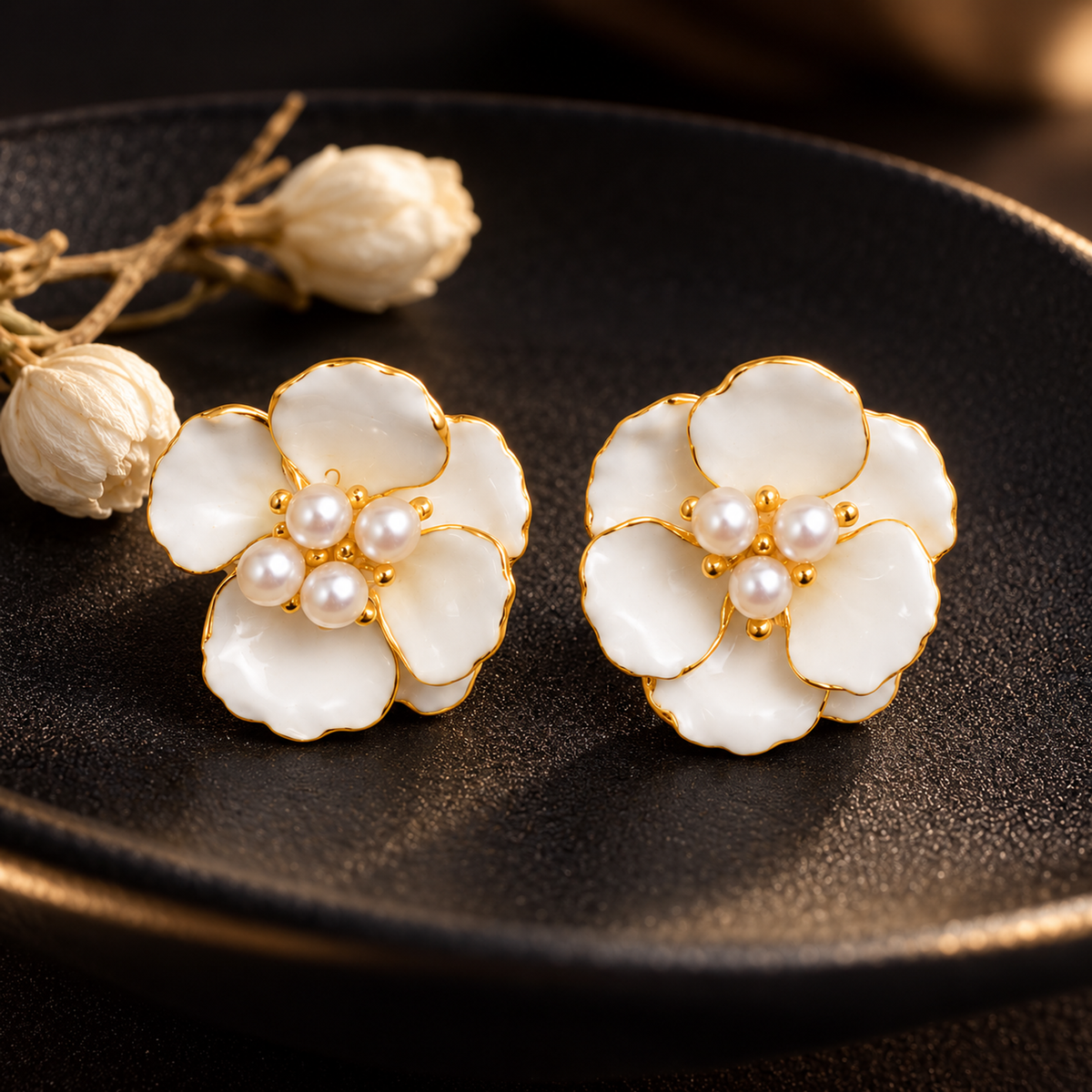 Two flower stud earrings on a minimalist matte black dish next to a small dried jasmine sprig under soft spotlight.