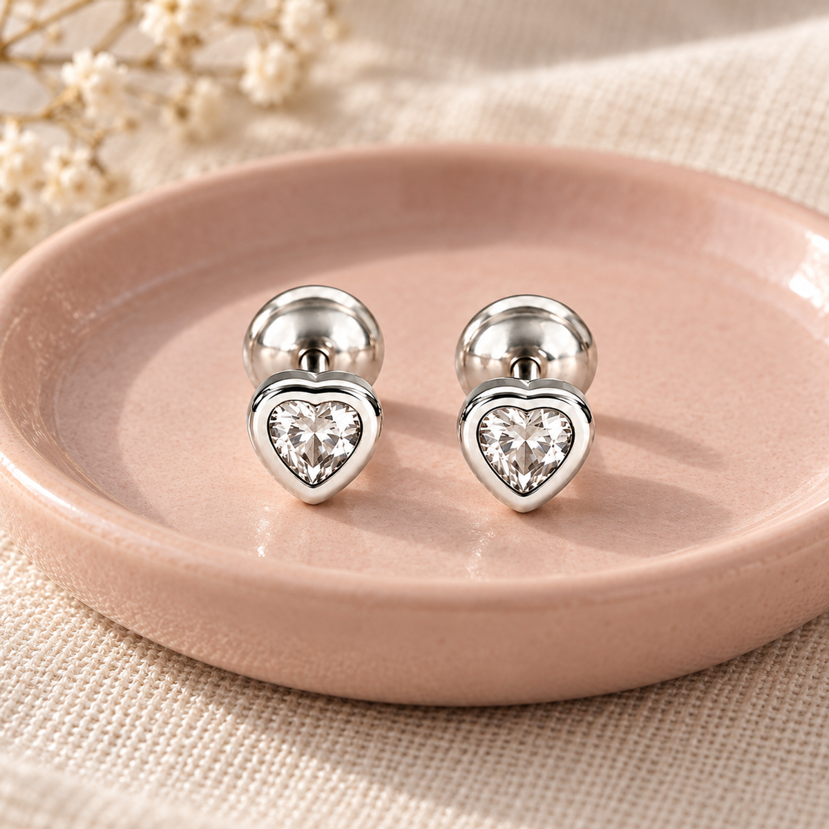 Silver heart stud earrings displayed on a blush pink ceramic dish over ivory linen.