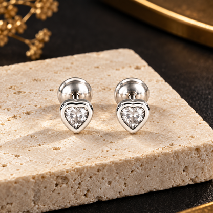 Silver heart stud earrings resting on an ivory stone pedestal against a matte black background.