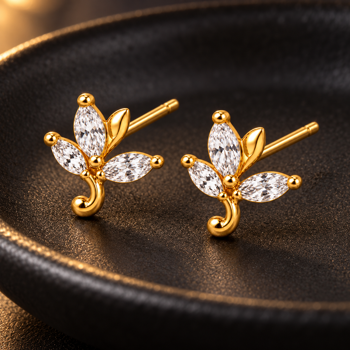 Gold leaf stud earrings on a textured matte black ceramic dish with warm lighting.