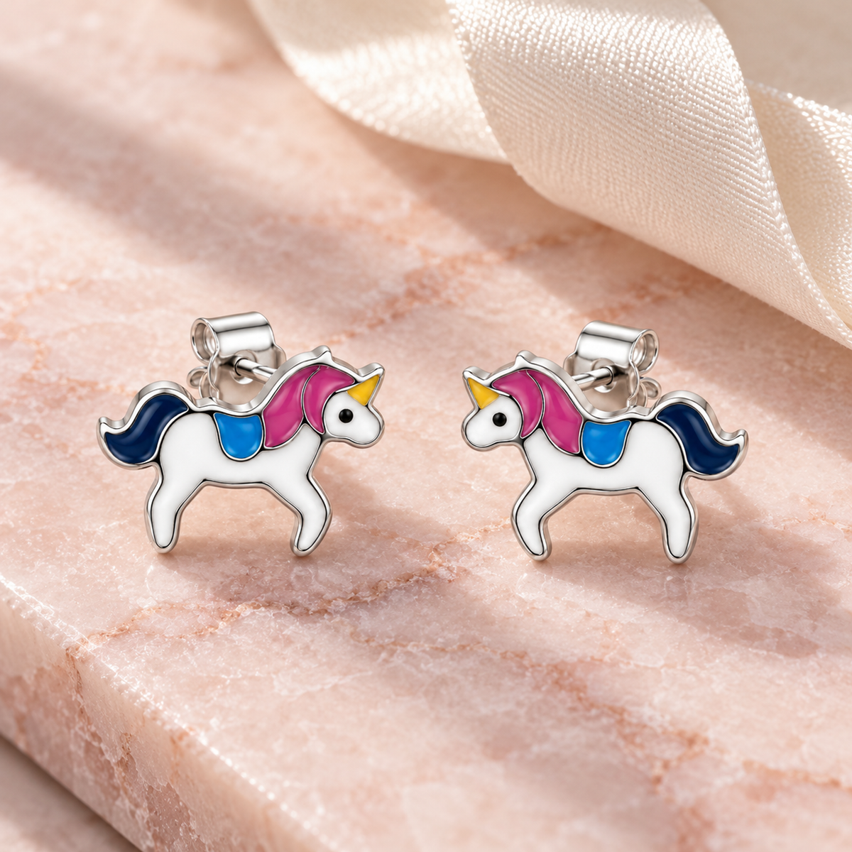 Silver unicorn stud earrings displayed on a blush pink marble slab beside an ivory silk ribbon.