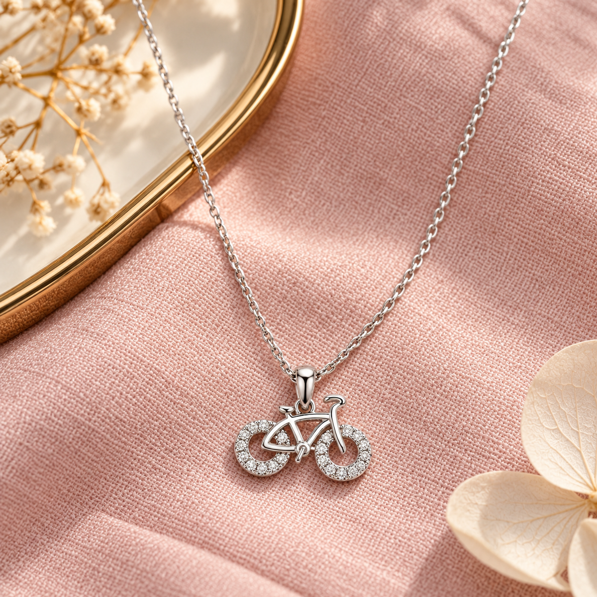 Silver bicycle necklace styled on blush pink linen alongside a warm gold tray.