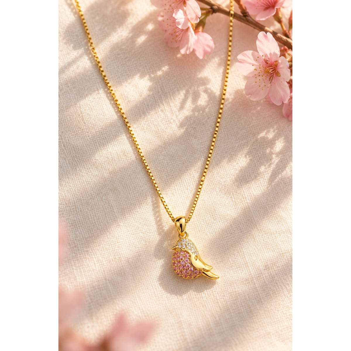 Gold bird pendant necklace arranged gracefully on ivory linen next to delicate pink cherry blossoms.