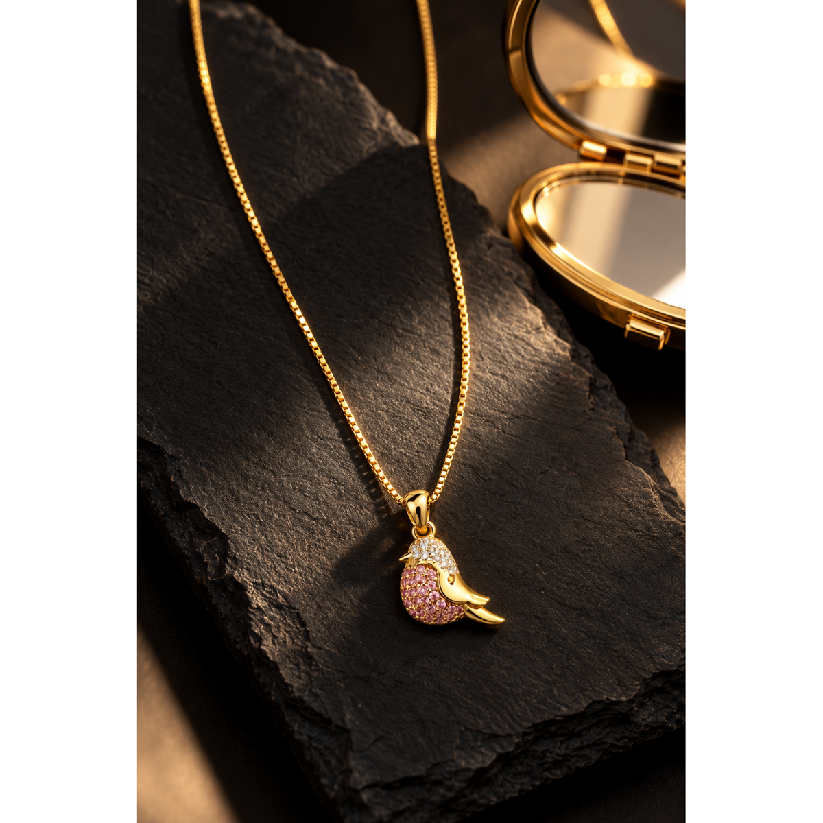 Sparkling gold bird pendant necklace presented on dark matte slate tile beside a gold compact.