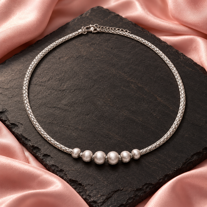 Woven silver bead choker laid on black slate next to blush pink silk fabric under soft studio light.