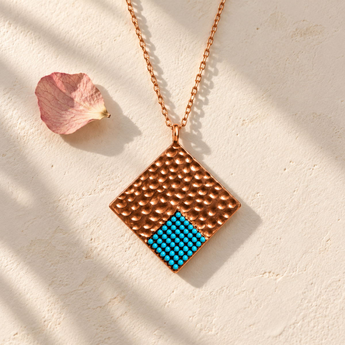 Rose gold geometric necklace lying on smooth ivory plaster beside a delicate dried petal.