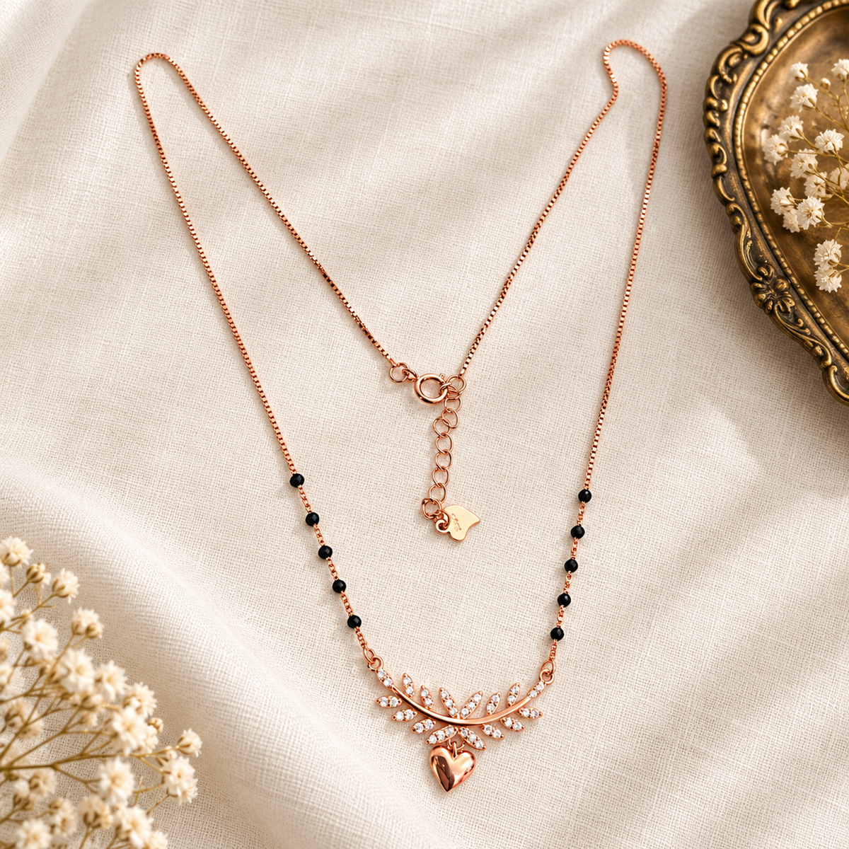 Rose gold mangalsutra displayed on textured ivory fabric beside brass accents.
