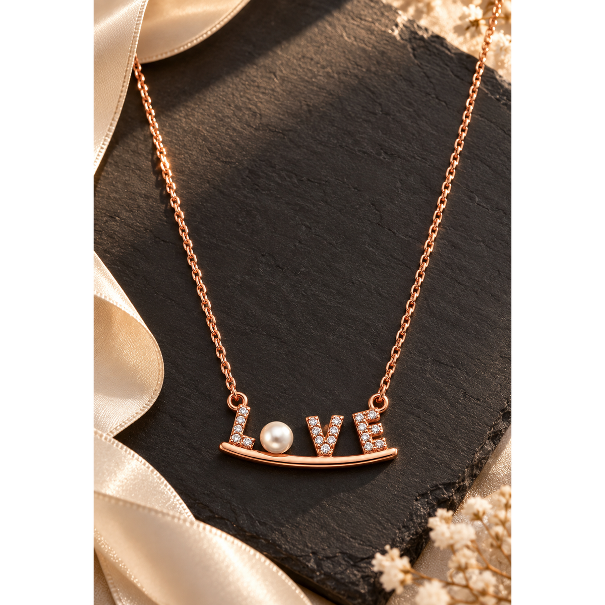 Rose gold LOVE necklace on a matte black stone surface accented by cream ribbon.