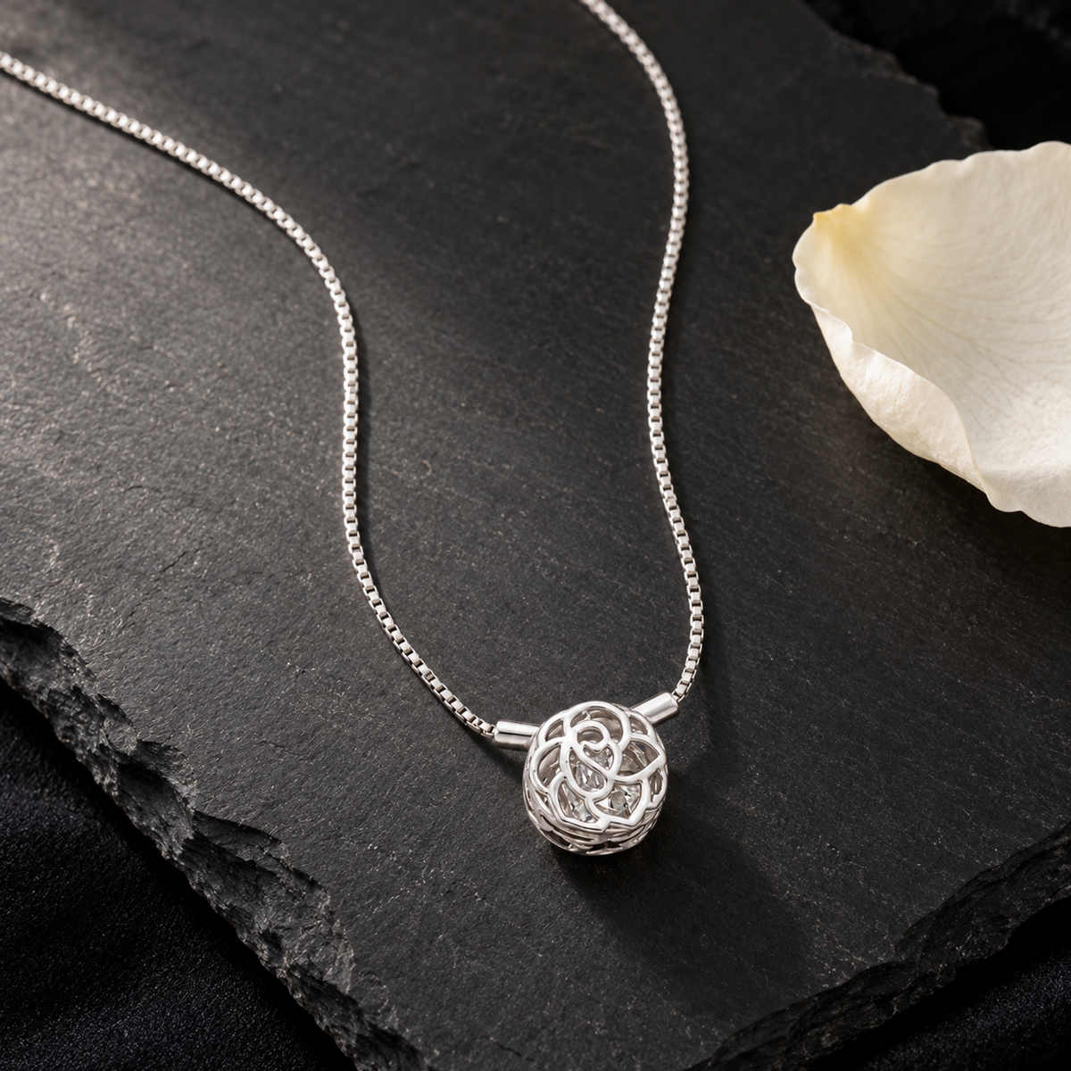 Delicate silver rose cage pendant necklace draped on a matte black slate tile next to an ivory rose petal.