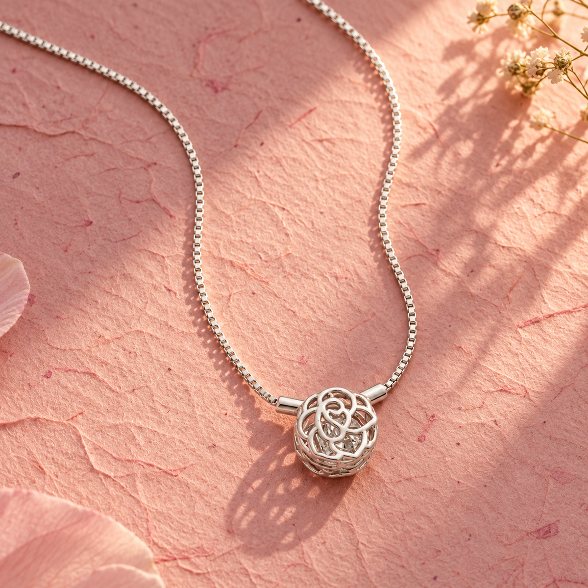 Silver rose cage necklace on textured blush pink paper under warm, angled golden hour light.