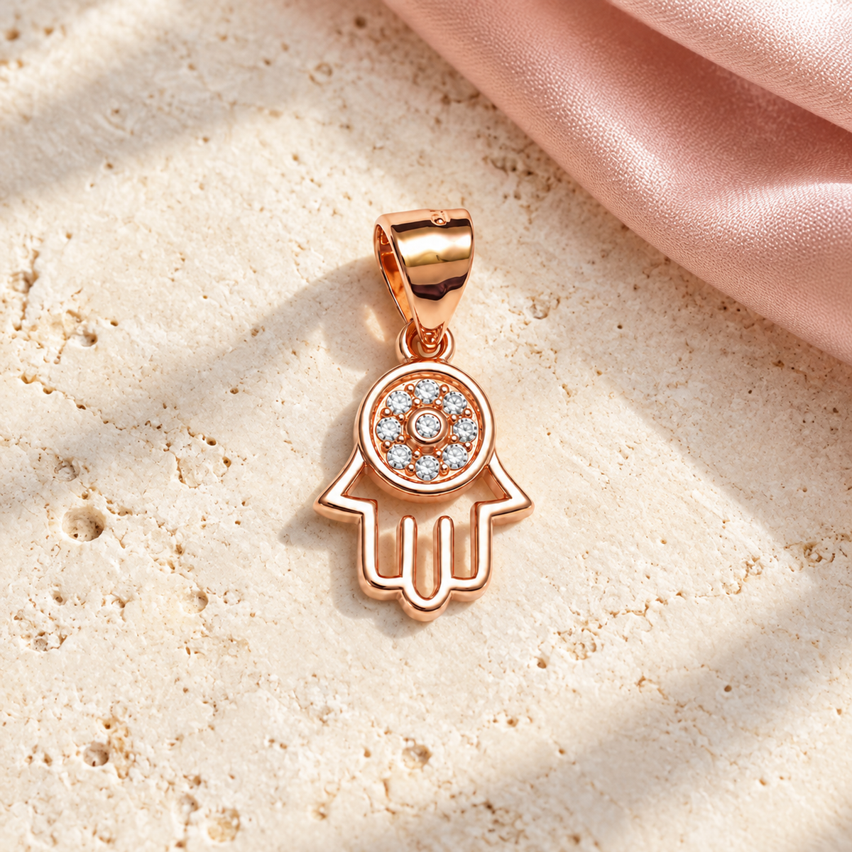 Rose gold Hamsa pendant resting on ivory stone alongside draped blush pink silk.