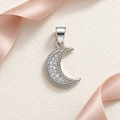Silver crescent moon pendant resting on ivory paper next to blush pink ribbon.