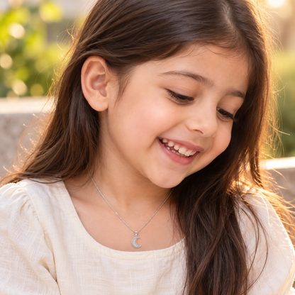 Child wearing a small silver crescent moon pendant on a chain over a white linen top.