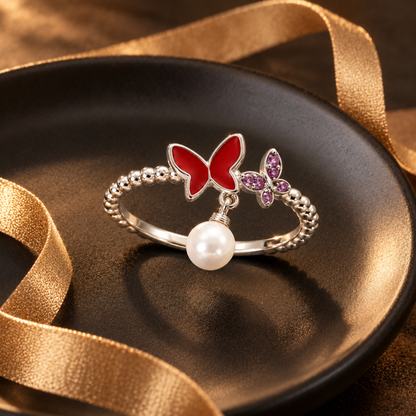 Silver butterfly ring with a dangling pearl on a matte black ceramic plate accented by a gold ribbon.