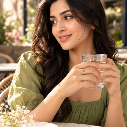 A woman wearing a silver enamel heart ring holds a glass, bathed in warm afternoon sunlight.