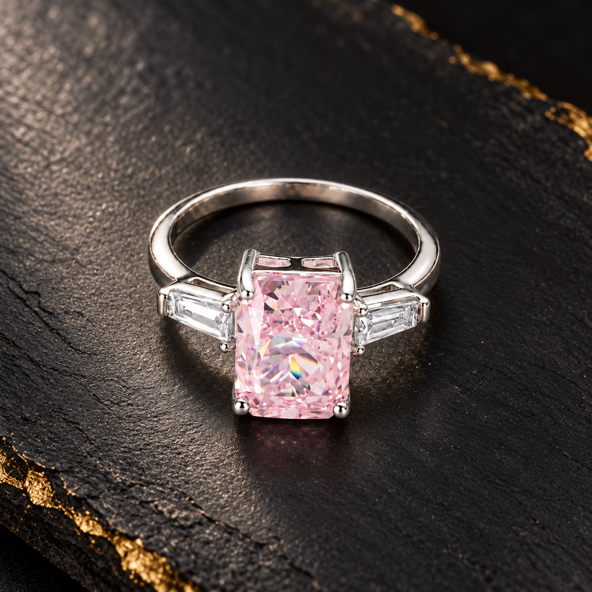 Overhead shot of a silver ring with a pink gemstone on a dark matte black slate with gold accents.
