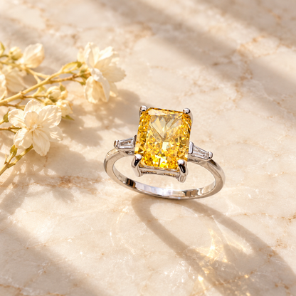 Overhead view of a silver ring with a yellow stone on ivory marble next to a dried jasmine sprig.