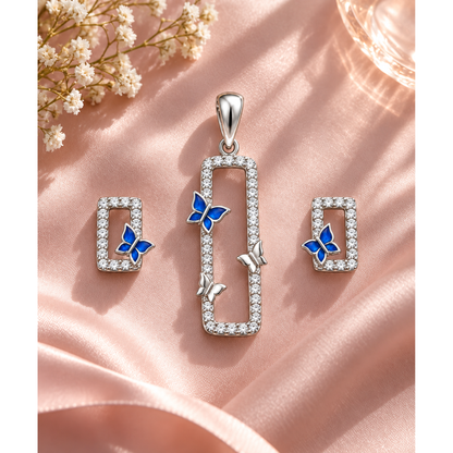 Silver pendant and stud earring set with blue butterfly detail laid on soft blush pink satin fabric.